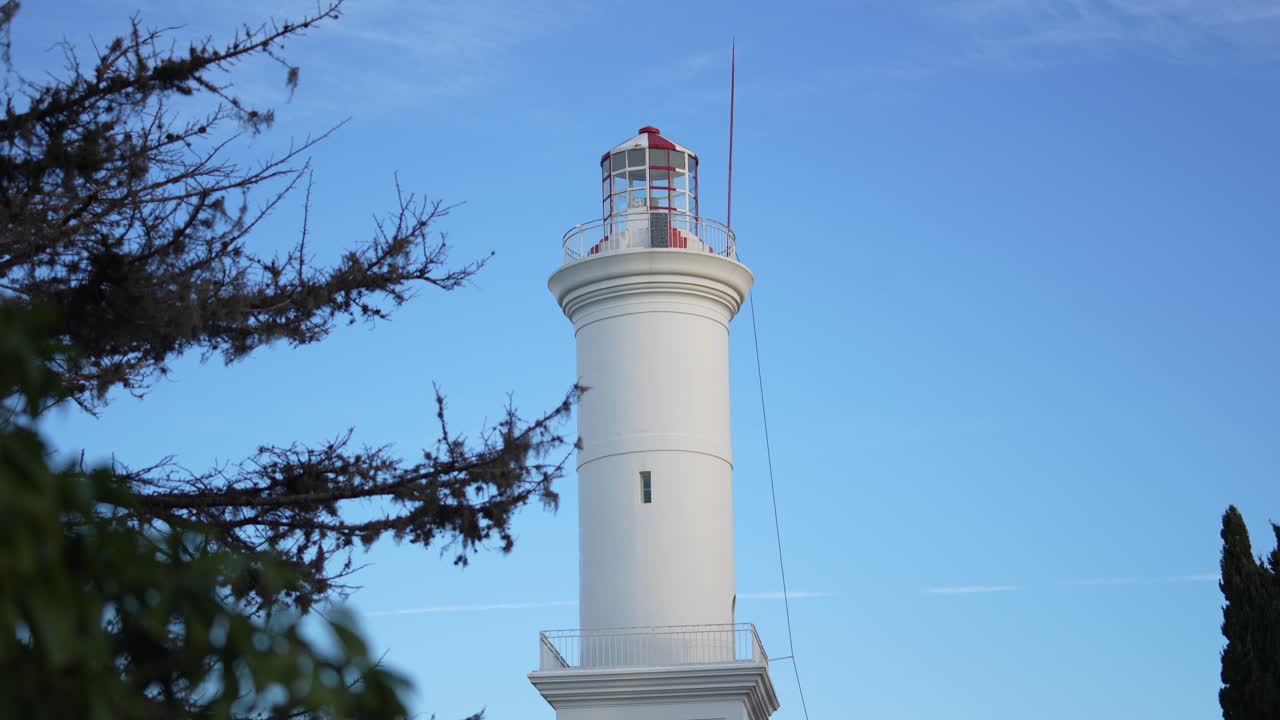 A close-up view of a white lighthouse against a clear blue sky, partially framed by tree branches.