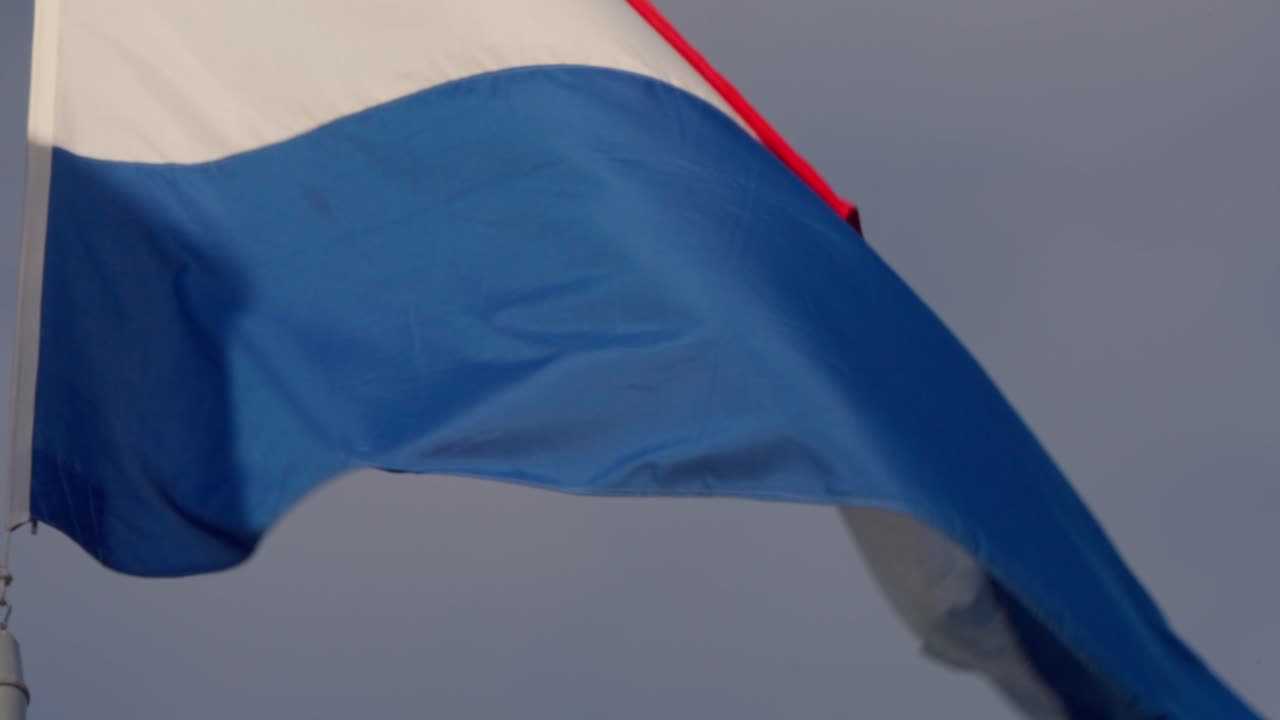 Red, white, and blue stripes of the Netherlands flag ripple in strong daylight breeze, filmed in Amsterdam, Netherlands (Nederland)
