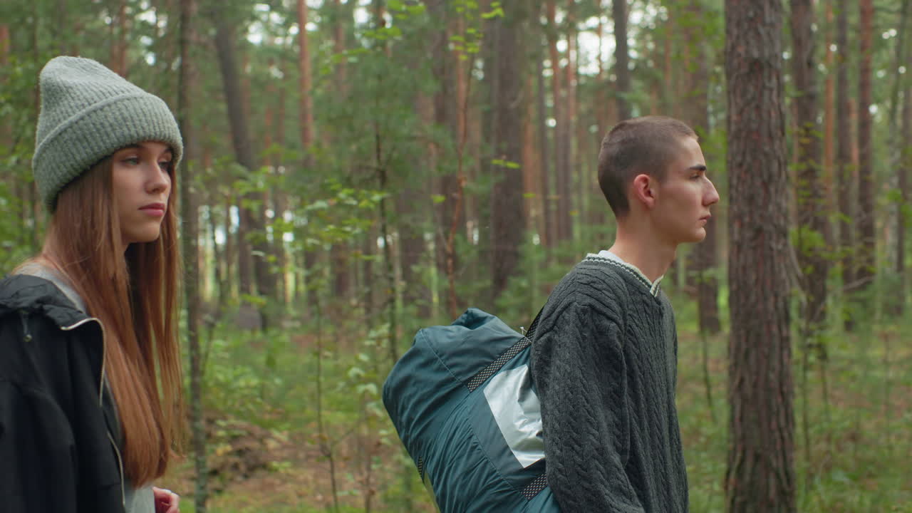Young travelers walk through dense forest surrounded by tall trees and greenery, carrying travel bags and observing nature with serious expressions, immersed in peaceful woodland