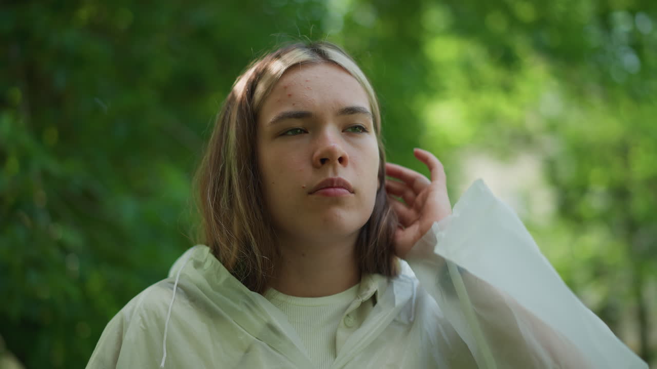 반투명한 레인코트를 입은 젊은 여성은 생각나게 바라보고, 평온하고 푸른 야외 환경에서 그녀의 자세를 조정하는 것처럼 보입니다. 그녀의 표정은 반사적이고, 생각에 빠져 있습니다.