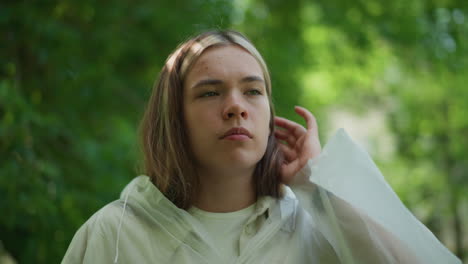 joven con impermeable translúcido mira pensativo, aparentemente ajustando su postura en un entorno al aire libre sereno y verde, su expresión es reflexiva, perdida en pensamientos