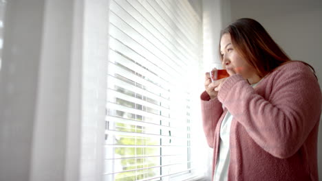 plus size biracial woman drinking tea and looking out of bedroom window, copy space, slow motion