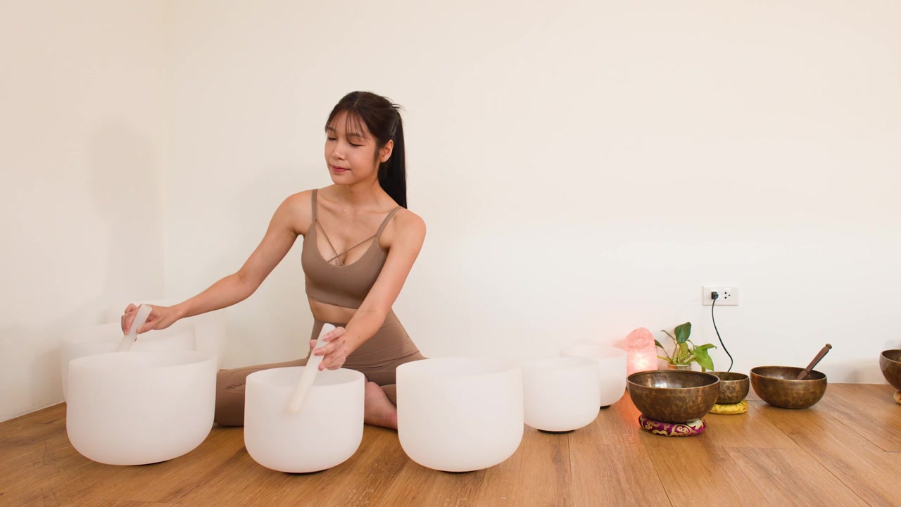 Femme asiatique en tenue de sport marron jouant des bols chantants en cristal pour la méditation dans un studio