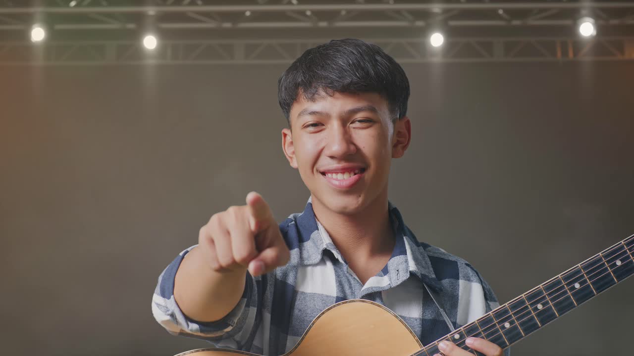 близкий взгляд на азиатского подростка с гитарой, делающего честный жест и улыбающегося на сцене.
