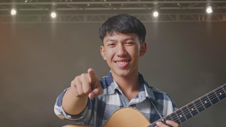 Primo piano di adolescente asiatico con la chitarra che fa un gesto di onestà e sorride sul palco.