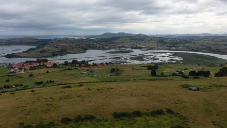 I-fly-with-a-drone-over-a-plateau-with-horses-grazing-and-we-see-the-mouth-of-a-river-to-the-sea-with-a-town-with-green-meadows-on-a-cloudy-day-in-summer-in-Cantabria-Spain