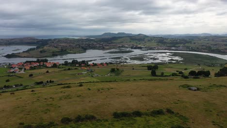 vuelo con un dron sobre una meseta con caballos pastando y vemos la desembocadura de un río al mar con una ciudad con prados verdes en un día nublado de verano en cantabria, españa