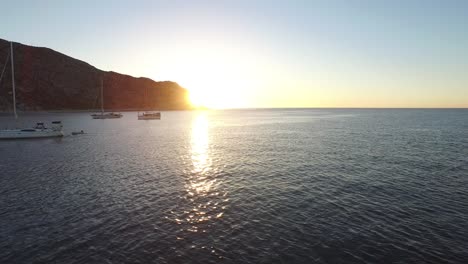 Toma-Aérea-De-Veleros-Y-Un-Yate-Con-El-Sol-Detrás-De-Una-Formación-Rocosa,-Parque-Nacional-Cabo-Pulmo,-Baja-California-Sur