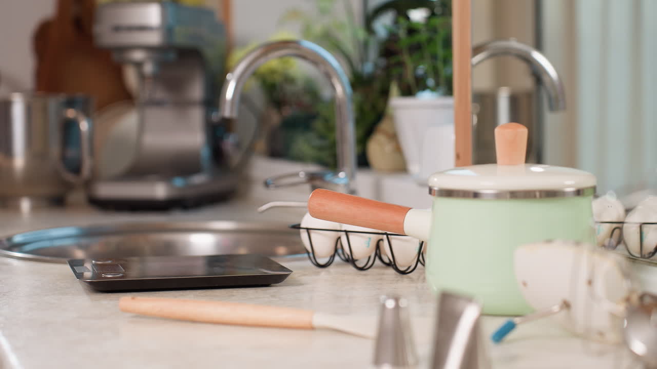 Modern kitchen setup with countertop showing mint green pot , wire egg rack with white eggs, stainless sink, digital kitchen scale, utensils, potted plants, and blurred mixer in background
