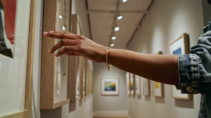 Mano destra di un visitatore che esamina una stampa lungo la galleria, con manica di jeans e braccialetto d'oro che sfiorano il vetro