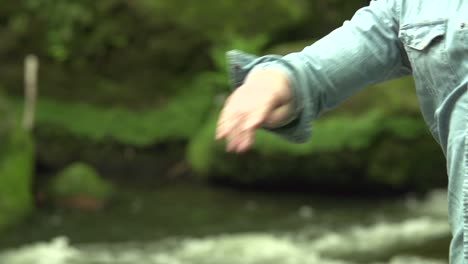 Mann-Wirft-Einen-Steinkiesel-Ins-Wasser-In-Der-Natur-Im-Wald-Mit-Wasser-Und-Steinen-Im-Hintergrund-In-Zeitlupe,-Nahaufnahme