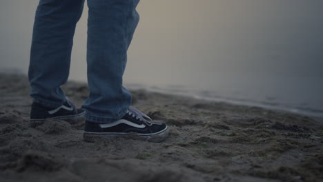 man legs in sneakers standing on sea shore. guy in blue jeans posing on beach