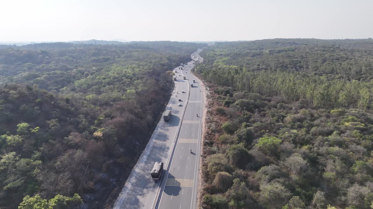 Aerial view of a multi-lane highway winding through dense green forests under a clear sky, with cars and trucks