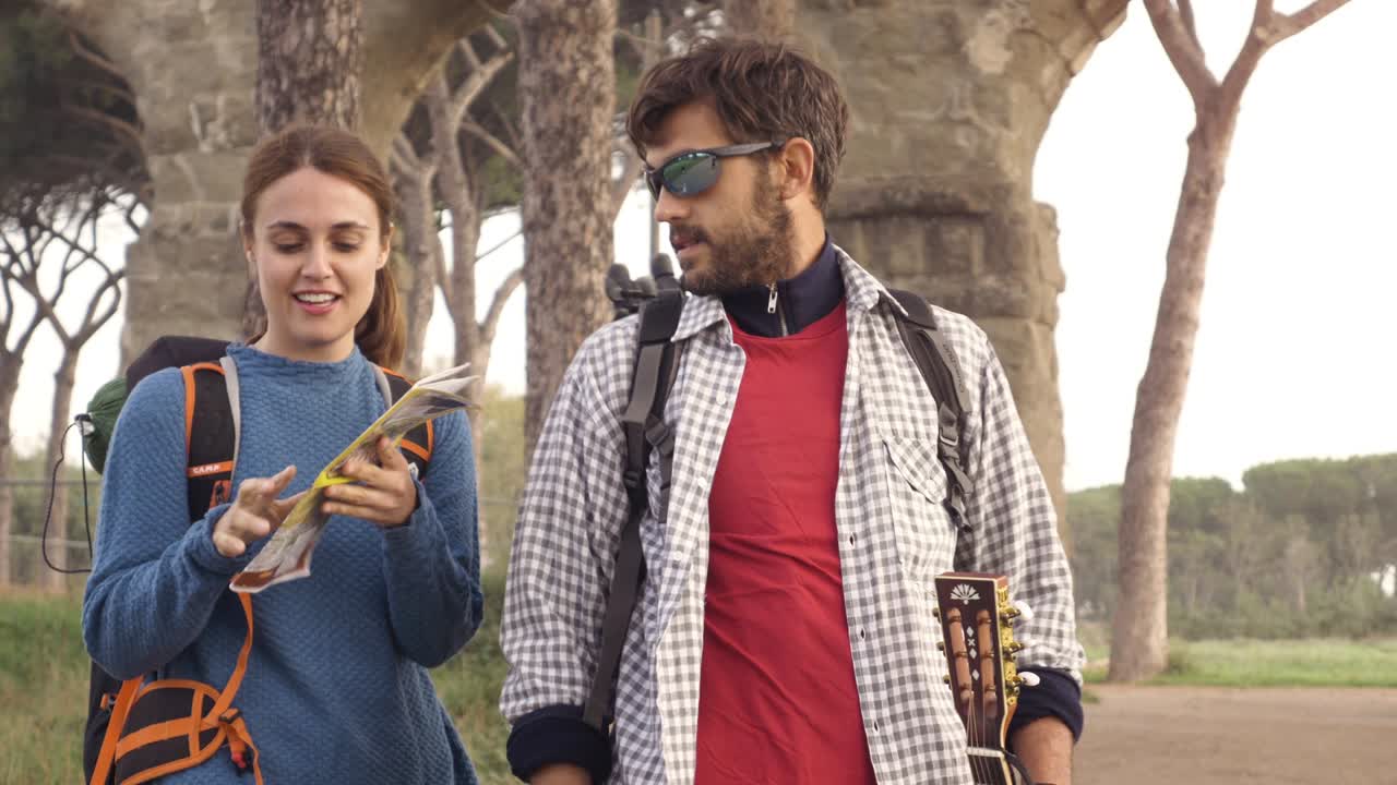 pareja feliz mochileros turistas que leen la guía del mapa para las direcciones perdidos cerca del acueducto romano arcos en el parco degli aquedotti ruinas del parque en roma al amanecer con guitarra y saco de dormir cámara estacionaria de cámara lenta
