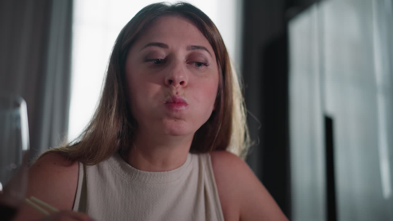 Young woman in sleeveless gown eating elegant cuisine with chopsticks, holding food close to lips, focused expression, indoor dining scene with soft lighting and moody background