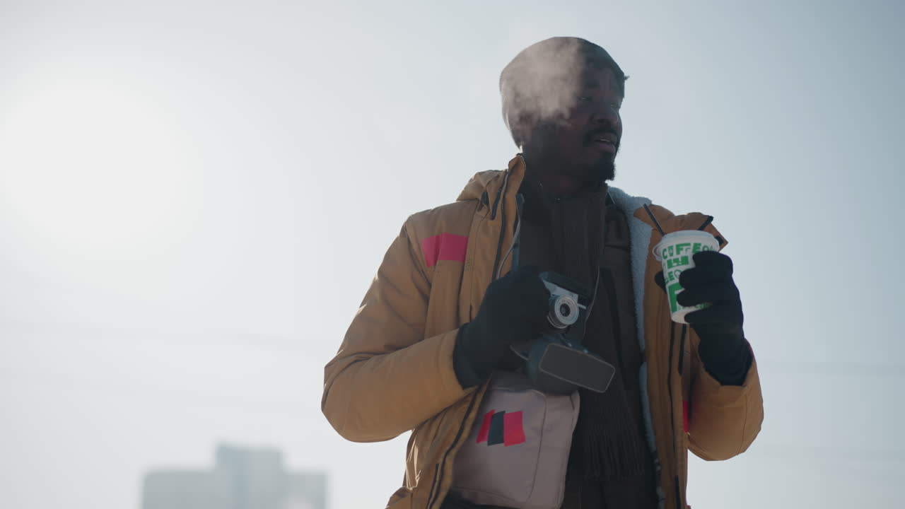 Cameraman di colore che indossa un berretto di lana e una giacca invernale, tiene caffè caldo e macchina fotografica, con una borsa a tracolla sul petto, che cammina per una strada urbana innevata sotto il sole splendente d'inverno, con il respiro visibile nell'aria fredda e frizzante.