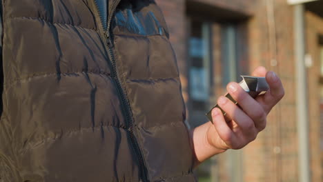 close up of person in padded vest holding cigarette pack while standing in front of building, fingers slowly closing cigarette pack, warm sunlight casting clear shadows on brick wall in background