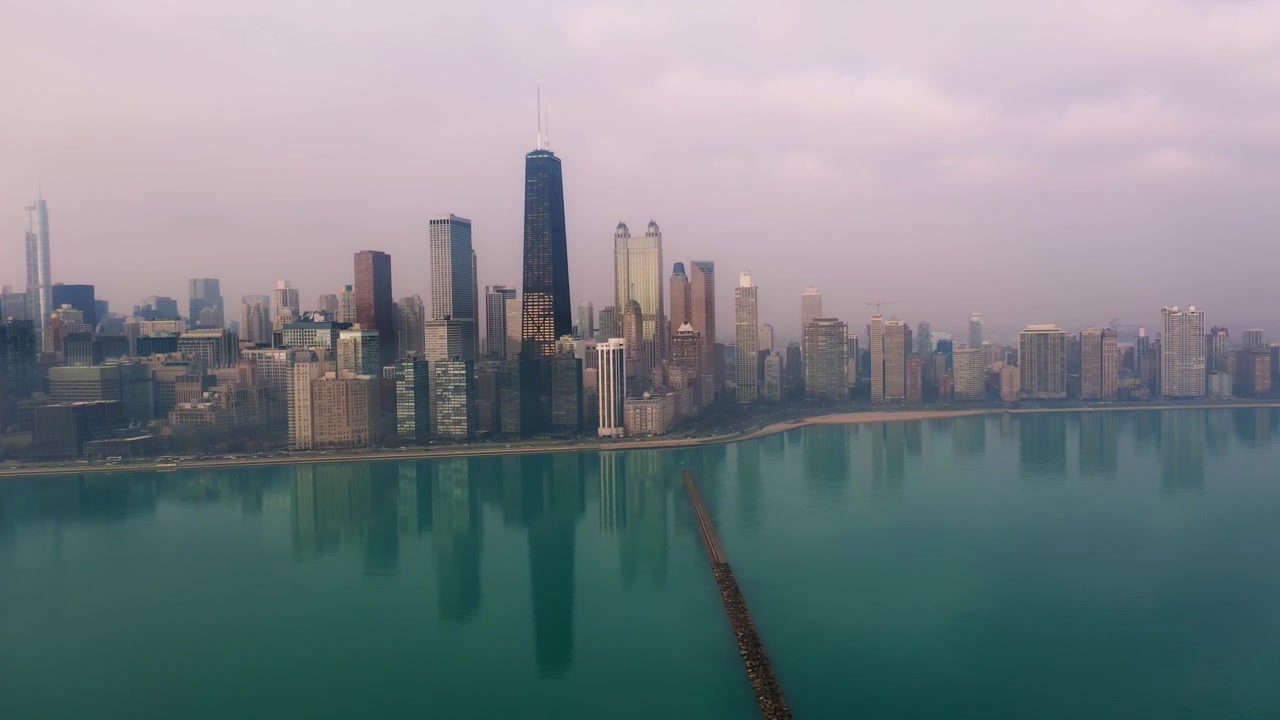 Smooth aerial footage pans right across Chicago on a hazy morning, showing the downtown skyline beside calm Lake Michigan with subtle reflections across the water.