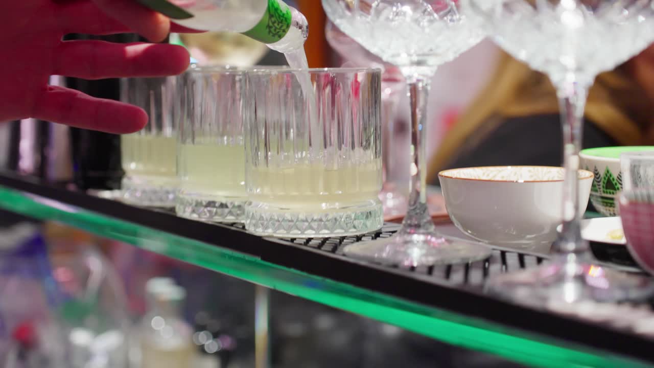 Bartender pouring a drink from a bottle into a glass at a bar counter with cocktail glasses