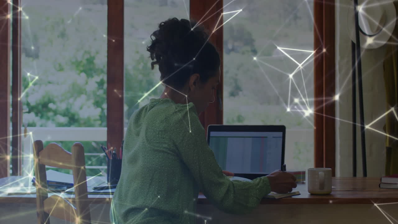 Mature woman referencing business spreadsheet on laptop while writing notebook with colored pencils