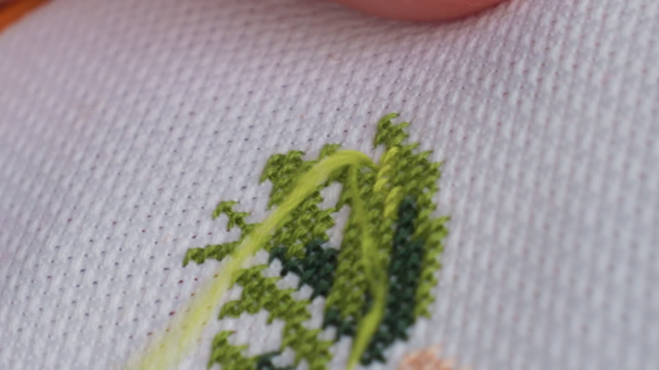 Ekstremt nærbillede af en dygtig håndværker, der fører en nål med grønt garn gennem et korsstingsstof, fingerspidser guider pæne x-mønstre, fine bevægelser, fokuseret udendørs håndarbejde i blødt dagslys