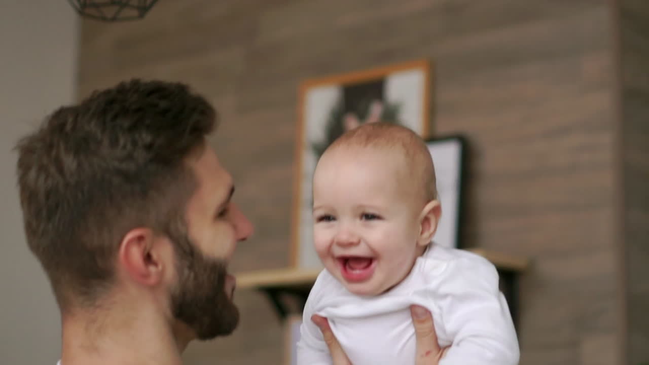 Father plays with infant son in white t-shirt looks at him and laughs. Laughing baby looking at the camera. Loving father