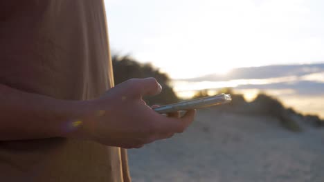 hombre joven sosteniendo el teléfono en la playa durante el atardecer