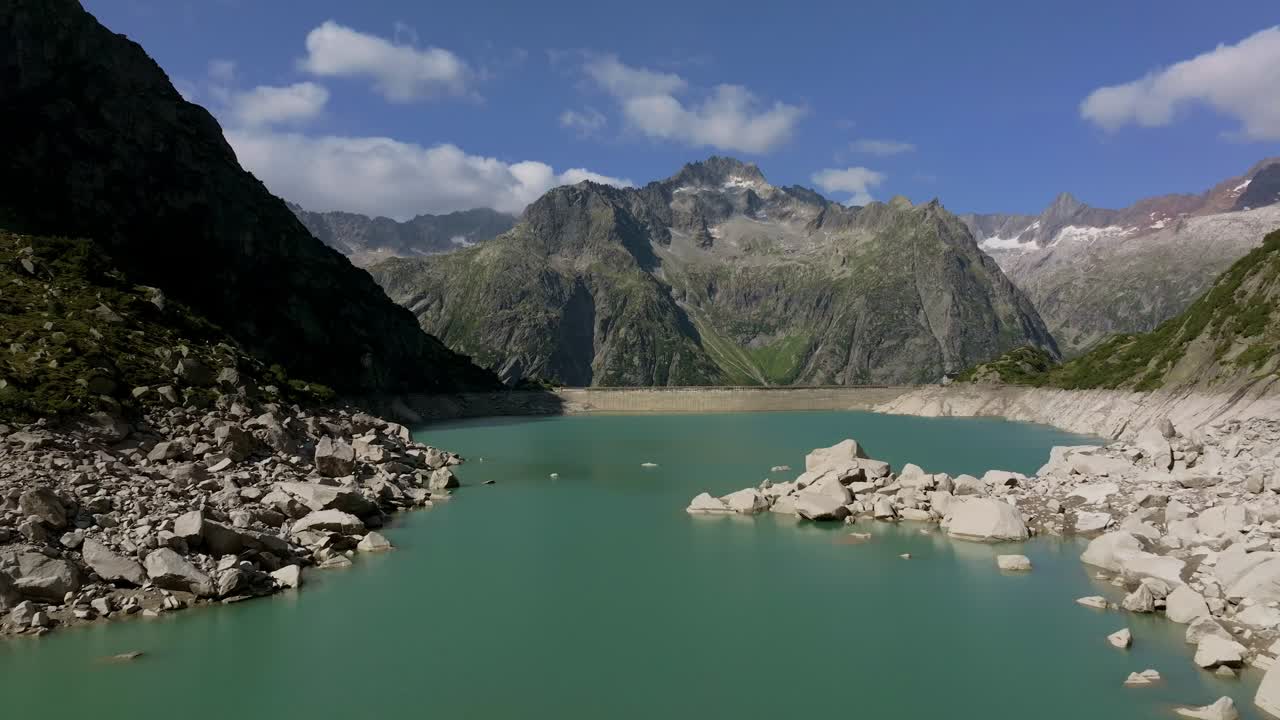 Aerial shot over the Gelmersee Lake in the Swiss Alps, featuring turquoise glacier-fed waters surrounded by steep granite peaks and rugged mountain terrain under a bright blue sky