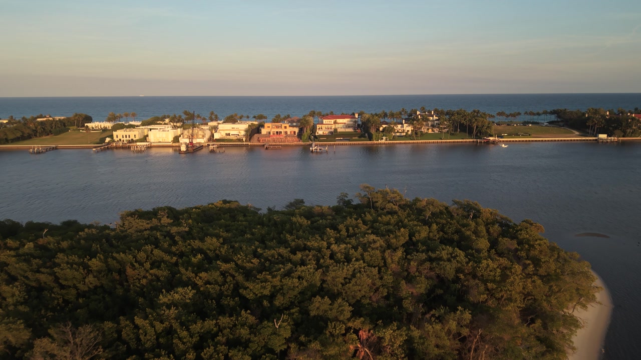 Drone in golden hour flying east over the ICW, from Summa Beach Park in West Palm Beach Florida toward the barrier island showing waterfront homes and the Atlantic Ocean in the distance.