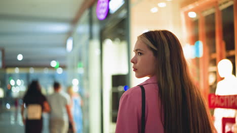 lady in pink dress walking through a mall, looking thoughtful with a blurred view of two people holding hands in the background, storefront mannequins are brightly lit, casting a warm glow