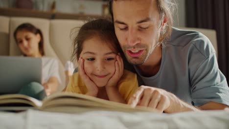 A-brunette-man-in-a-gray-t-shirt-is-reading-a-book-with-his-interested-little-brunette-daughter-in-a-yellow-t-shirt,-while-her-mother-is-relaxing-in-the-background-and-lying-on-the-sofa-with-a-laptop-in-a-modern-apartment