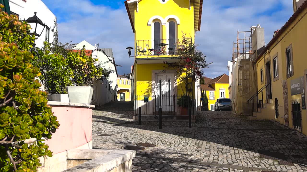 Vista dal basso di un'affascinante casa gialla nella tipica via portoghese con facciate colorate, pavimentazione classica in ciottoli di calçada e piante in vaso in una tranquilla e soleggiata atmosfera da piccola città