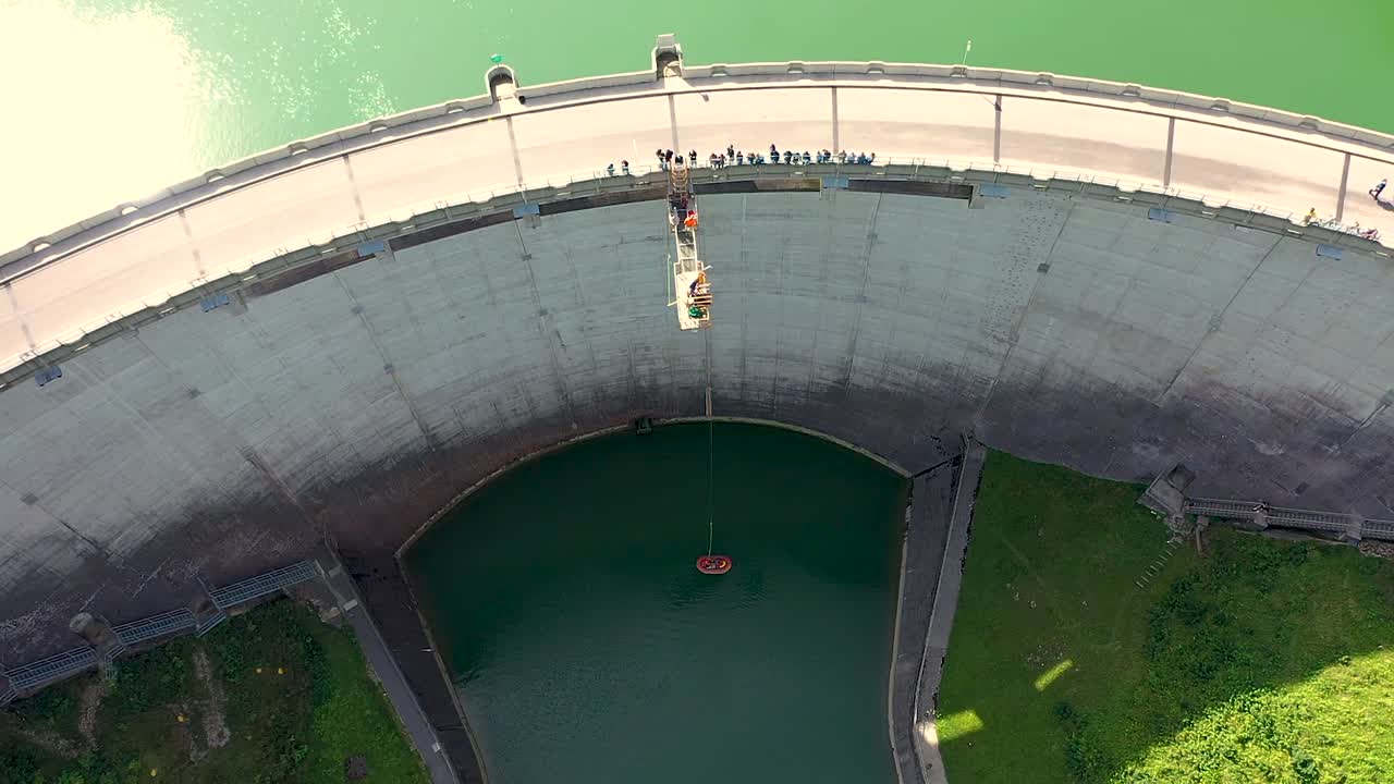 Birds eye top down Aerial view of young people bungee jumping off a 50m high dam platform while viewers standing on dam bridge watching action happen