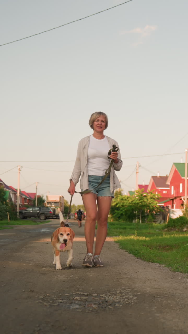 mujer caminando con su perro en la correa a lo largo de una carretera rural, el fondo con edificios de colores, vegetación y charcos en el camino de grava, mostrando una escena al aire libre relajada