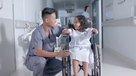 diverse male nurse and child patient sitting in wheelchair in corridor at hospital, in slow motion