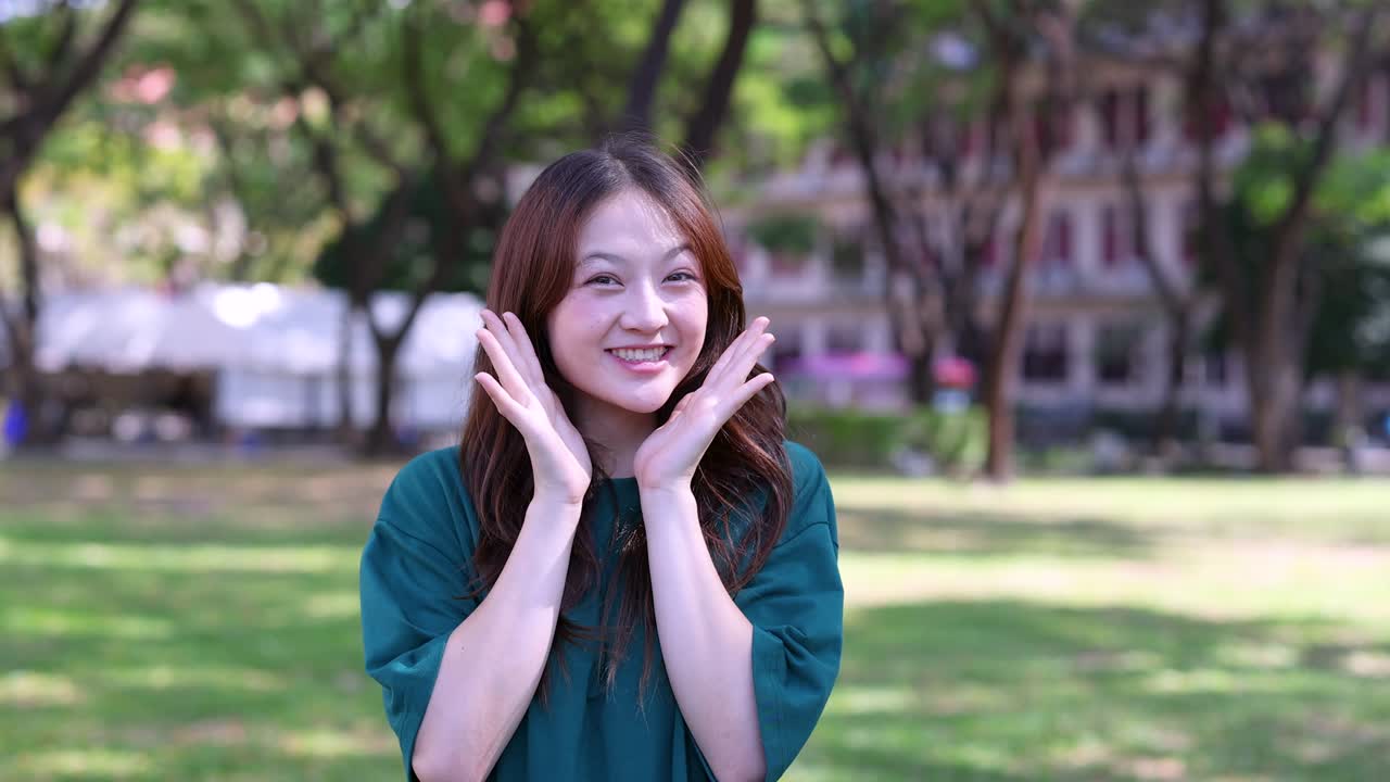 หญิงเอเชียอายุน้อยแสดงอาการตกใจและตื่นเต้นขณะยืนอยู่ในสวนที่มีแสงแดดส่องถึง