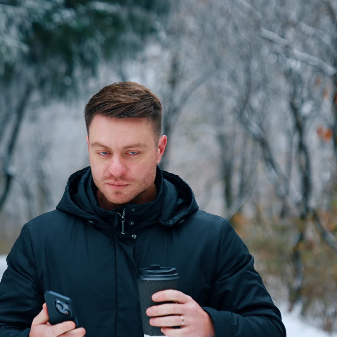 Man wearing black jacket stands in the winter park. Caucasian male drinks coffee and looks at his smartphone