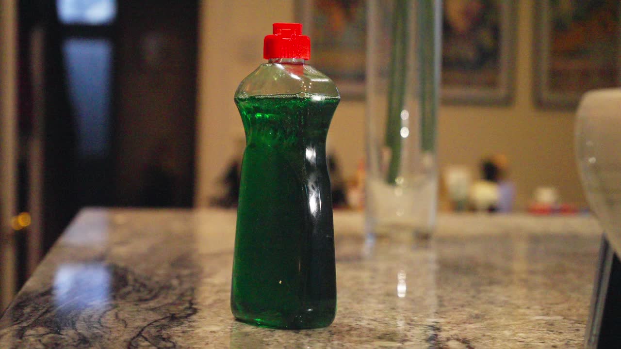 Close-up shot shows hand reaching for green dish soap bottle on kitchen counter