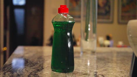 close-up shot shows hand reaching for green dish soap bottle on kitchen counter