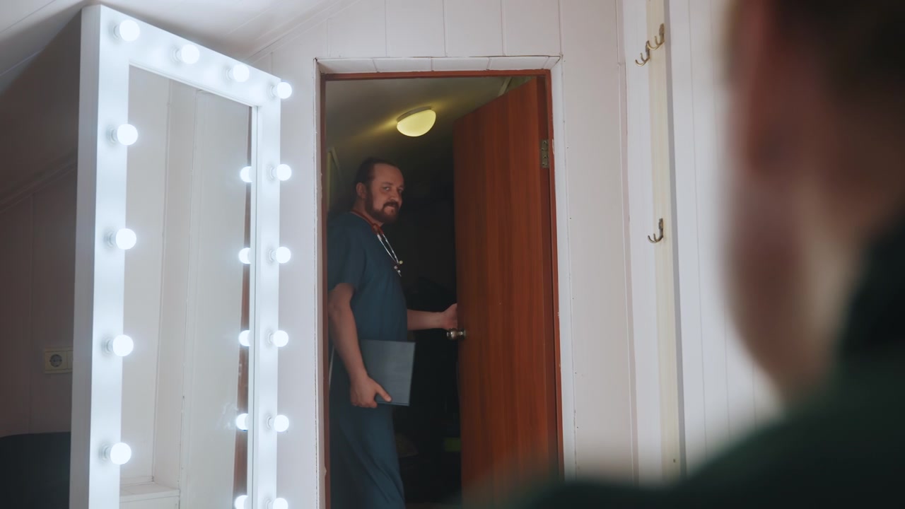 Paramedico che lascia la porta della mansarda; assistente maschio bianco cammina verso una porta chiusa accanto a uno specchio da trucco illuminato, passi lenti e misurati, ambiente domestico intimo, tensione contenuta, gesto di addio.