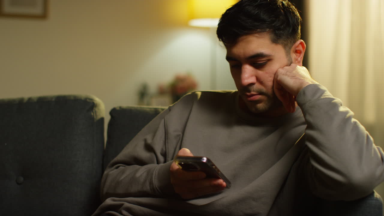 joven que pasa la noche en casa sentado en el sofá con el teléfono móvil desplazándose por internet o medios sociales transmitiendo contenido 4