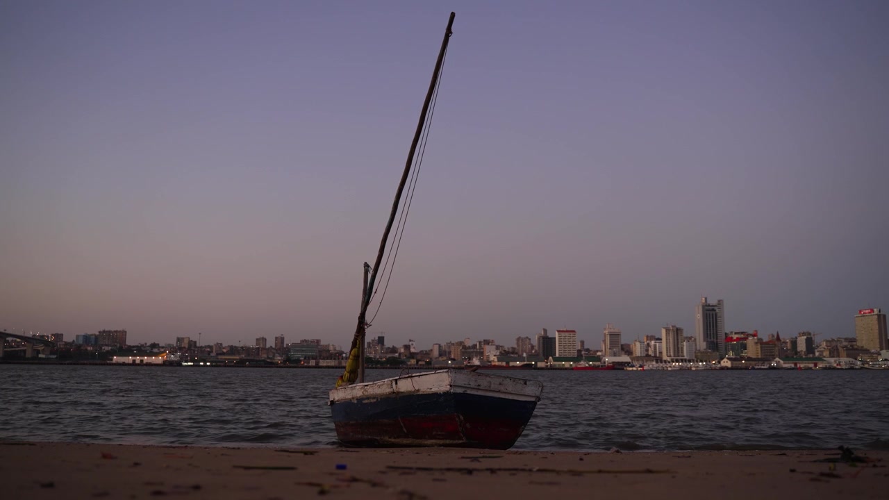 Una piccola barca da pesca arenata sulla sabbia lungo il lungomare con lo skyline della città di Maputo sullo sfondo e la luce soffusa del tramonto che conferisce all'aria un bagliore maestoso.