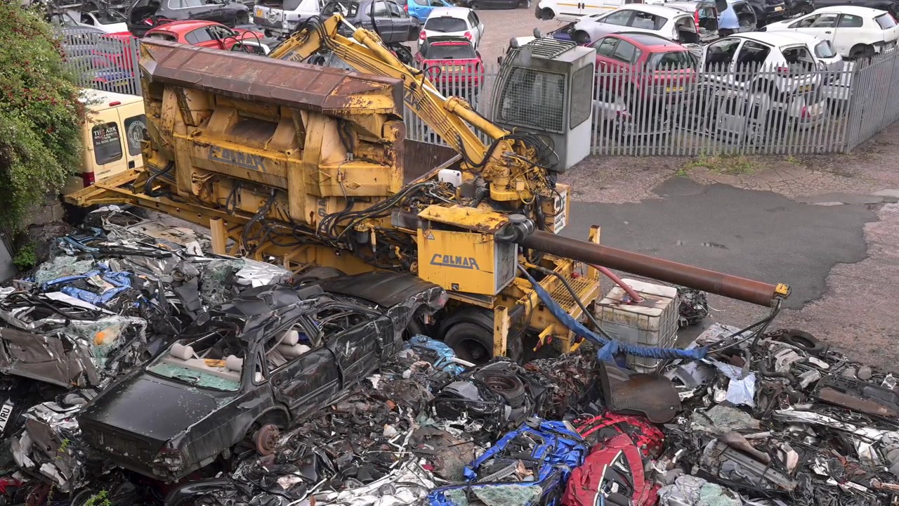Industrial car crusher works through piles of wrecked vehicles in a busy automotive scrap yard, viewed from above.