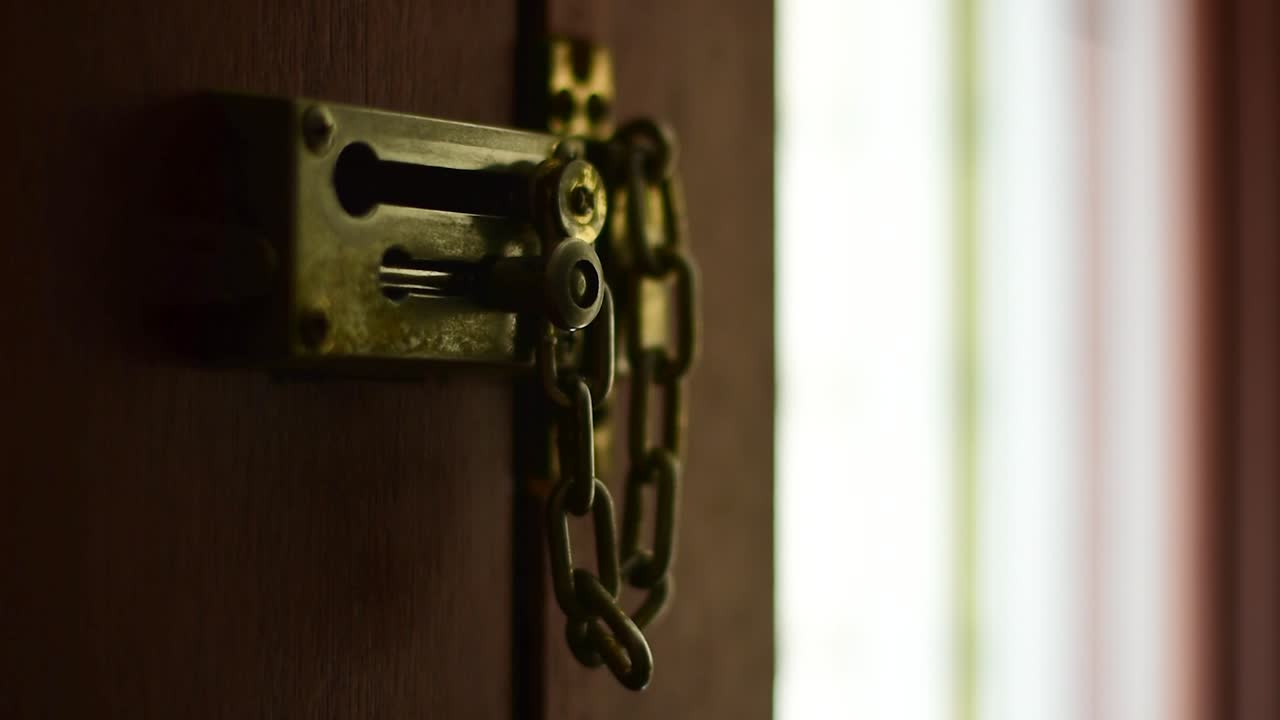 bellamente filmado de cerca de un cerrojo y una cadena desbloqueados en el interior de una puerta residencial, con una mano deslizando el cerrojo en su lugar y enganchando la cadena, con un elegante bokeh