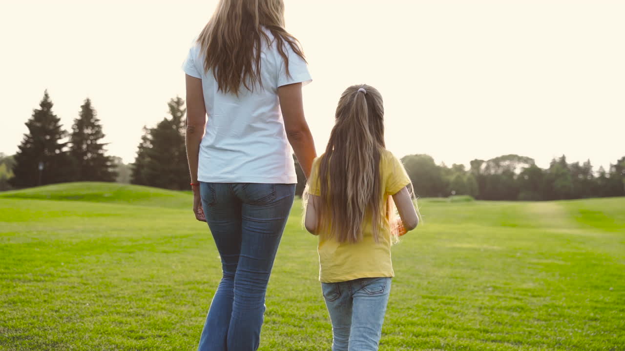 Free stock video - Back view of mother and little daughter holding hands and walking on green grass field in the park