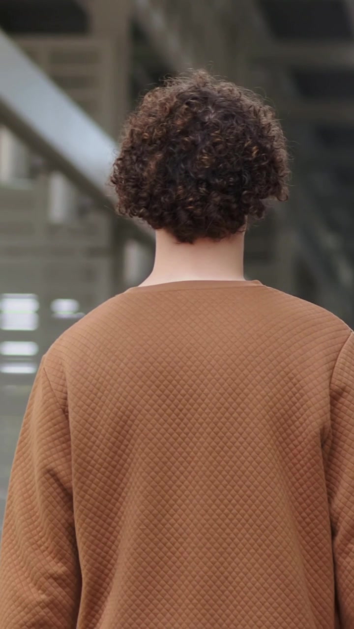 Vertical video: Starting walking person moving right inside large hall, with rust sweater, louvers