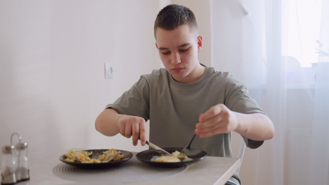 Teen boy in green shirt sits at dining table using fork and knife to eat scrambled eggs from black plate while focused in bright room with light curtains and peaceful morning atmosphere