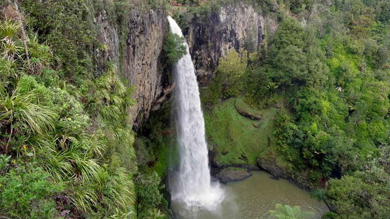 Looking at the huge Bridal Veil Falls waterfall from above, cliffs, trees and swimming hole in paradise on walking trail in Raglan, New Zealand Aotearoa