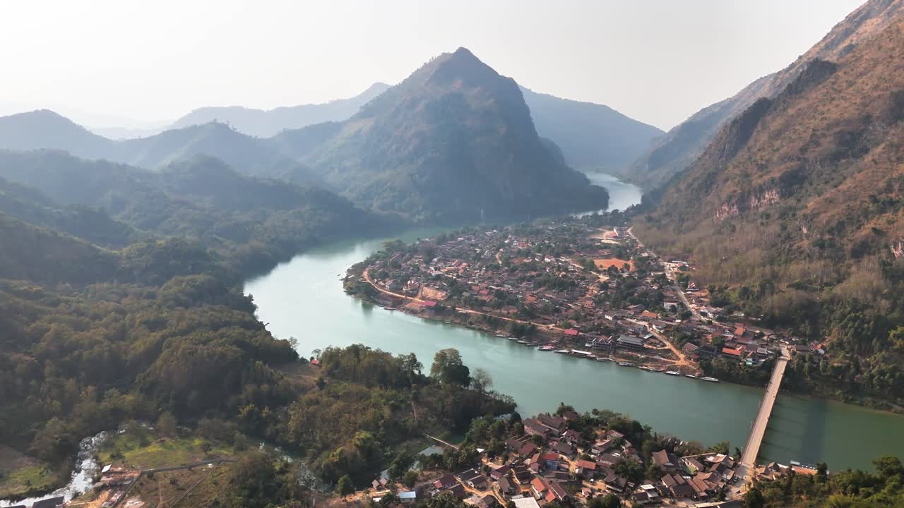 Aerial view of Nong Khiaw, Laos, with a scenic river winding through a lush mountain valley, featuring a village, a connecting bridge, and dramatic peaks under hazy daylight in a tranquil setting