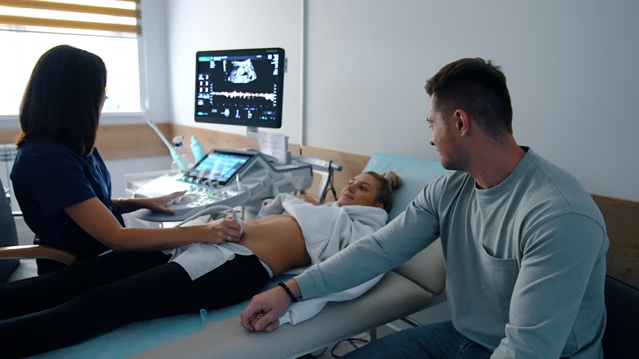 Happy future parents look at each other smiling seeing their baby image on the screen. Pregnant woman is checked on ultrasound machine.